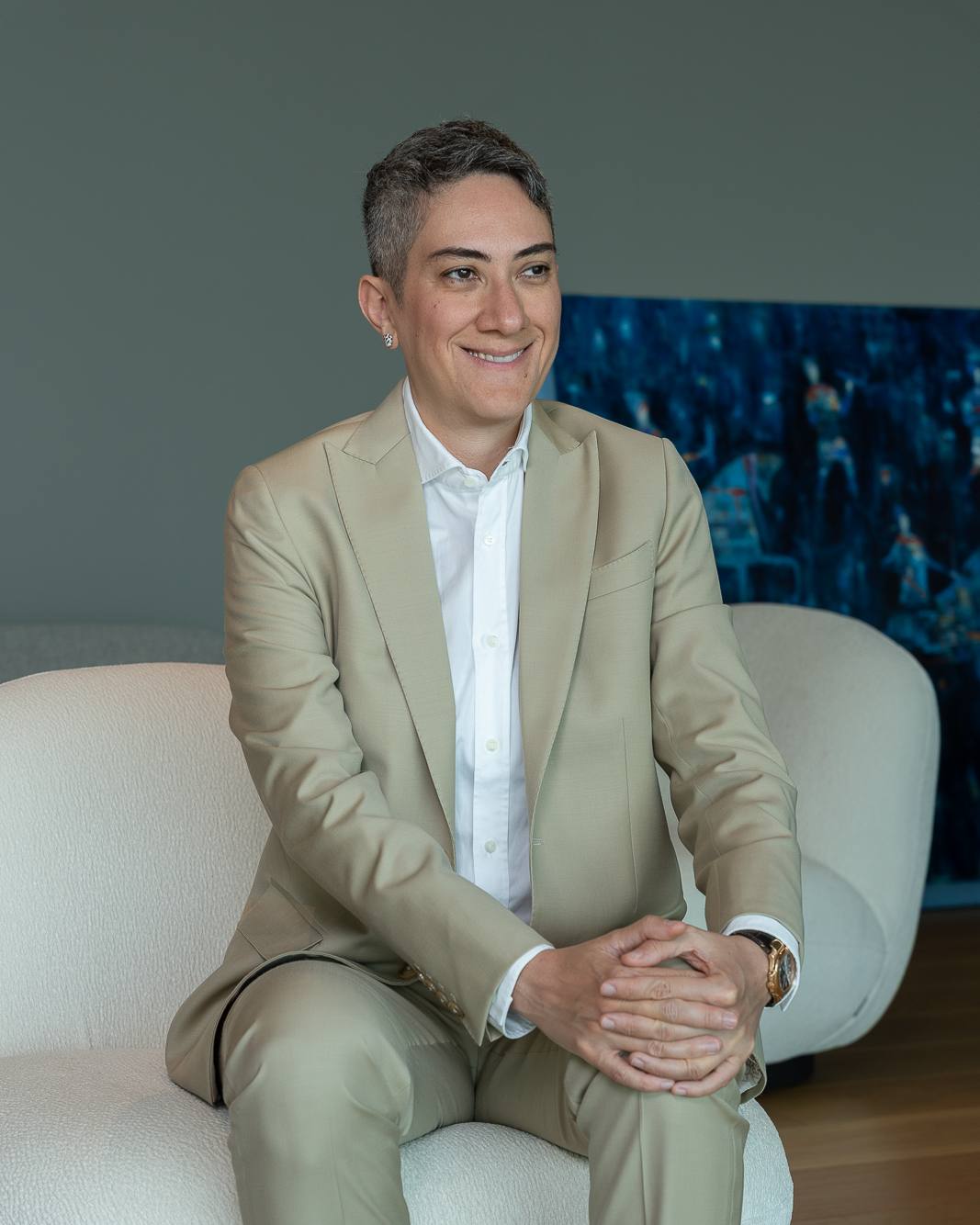 Honey Deylami laughing, in a beige suit, seated in a white armchair